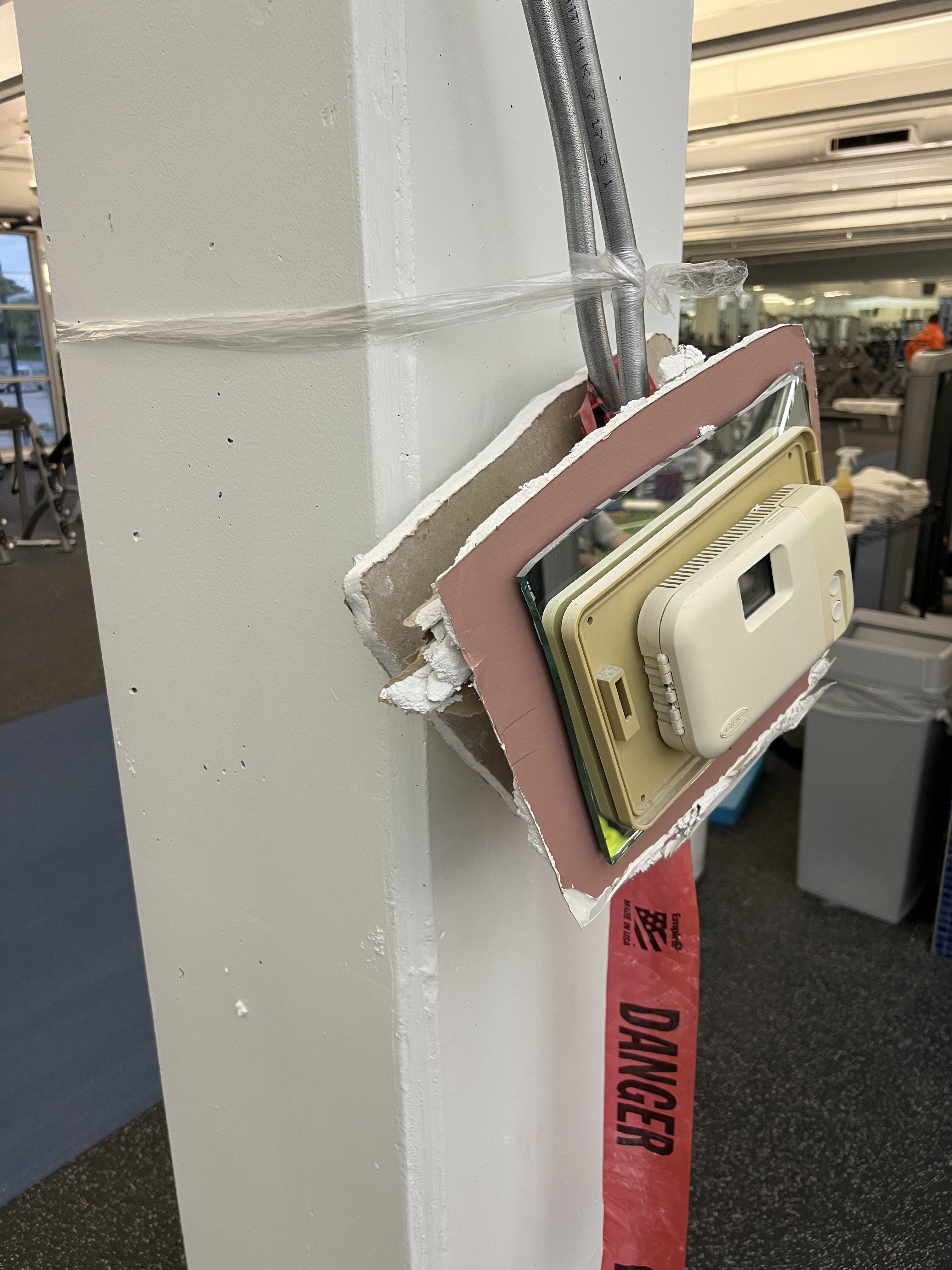 A thermostat hanging from a cut piece of mirror stuck to a cut piece of drywall hanging from a pillar in a public gym